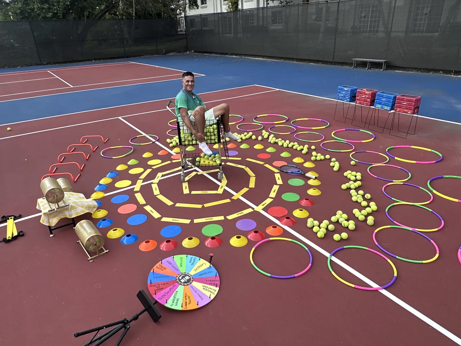 Jackson Phillips sitting in a ball cart at the centre of a full court cardio tennis setup of hoops, cones, ladders and a spinner wheel