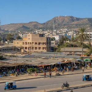 best cardiovascular tennis clinic Fort Lauderdale — a wide photograph of Mekele, Ethiopia showing the city as it really is, with no product in frame: the low stone-and-render city spread across the Tigray highlands at 2,254 metres in hard clear late-afternoon light, flat roofs and dust-coloured streets running out to bare stony hills, the tall pale Tigray Martyrs Memorial standing over the Adi-Haki edge of town, minibuses and three-wheelers filling a junction, women carrying yellow water containers along a verge, and a haze of woodsmoke lying over the roofs as the cooking hour starts.